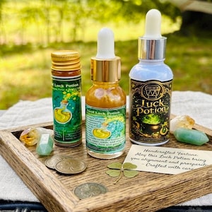 May include: Three glass bottles labeled Good Luck Potion, two green crystals, three bronze coins, and a four-leaf clover on a wooden tray. One bottle has a gold cap, one has a white dropper, and one has a silver dropper. A card reads Handcrafted Fortune.