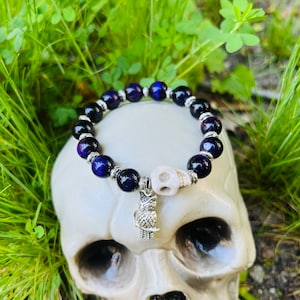 May include: A dark blue beaded bracelet with silver accents and a silver owl charm. The bracelet is displayed on a white skull. The beads are a mix of dark blue and black. The owl charm is detailed and adds a unique touch.