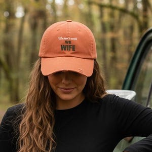 May include: An orange baseball cap with the text "We don't need no wife" in black lettering. The cap is distressed. The person is wearing a black long-sleeve shirt and camouflage trousers.