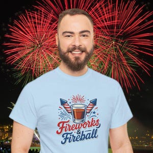 Może przedstawiać: Jasnoniebieska koszulka z napisem "Fireworks & Fireball". Grafika zawiera kieliszek, fajerwerki i rakiety w kolorach czerwonym, białym i niebieskim. W tle uśmiechnięty mężczyzna na tle pokazu fajerwerk&oacute;w.