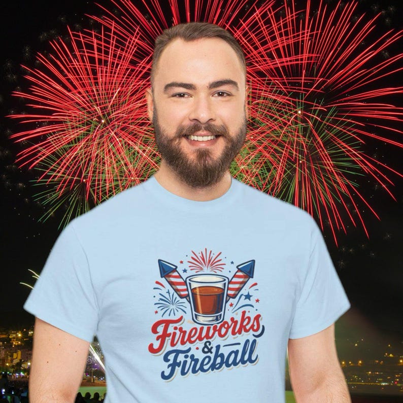 Może przedstawiać: Jasnoniebieska koszulka z napisem "Fireworks & Fireball". Grafika zawiera kieliszek, fajerwerki i rakiety w kolorach czerwonym, białym i niebieskim. W tle uśmiechnięty mężczyzna na tle pokazu fajerwerk&oacute;w.