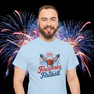 Może przedstawiać: Jasnoniebieska koszulka z napisem "Fireworks & Fireball" w kolorze czerwonym i niebieskim. Grafika zawiera drinka, fajerwerki i amerykańskie flagi. Tło przedstawia kolorowe fajerwerki na czarnym tle.