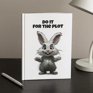 May include: A white journal with the text "DO IT FOR THE PLOT" and a cartoon illustration of a smiling gray bunny. The journal is on a dark surface with a pen and a lamp in the background.