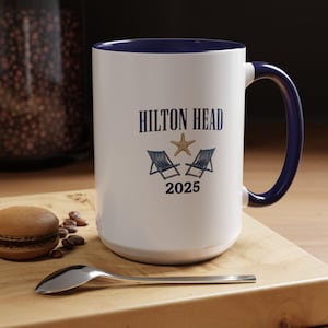 May include: White ceramic mug with a navy blue interior and handle. The mug features the text "HILTON HEAD" above a starfish and two beach chairs, with "2025" below. The mug is on a wooden surface with a macaron and spoon.