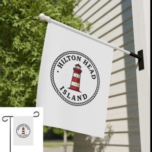 May include: White flag with a circular design featuring a red and white striped lighthouse, the words "HILTON HEAD ISLAND" in a black font, and a rope border. The flag is mounted on a white pole and black bracket.