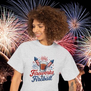 Może przedstawiać: Jasnoszara koszulka z grafiką z napisem "Fireworks & Fireball" w kolorze czerwonym i niebieskim. Grafika zawiera szklankę bursztynowego płynu, dwie rakiety i fajerwerki. Tło przedstawia pokaz sztucznych ogni.