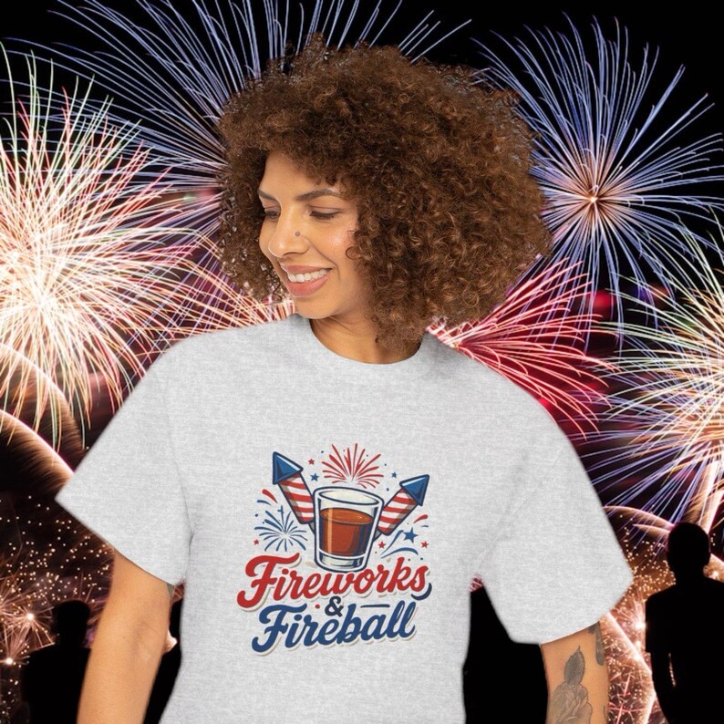 Może przedstawiać: Jasnoszara koszulka z grafiką z napisem "Fireworks & Fireball" w kolorze czerwonym i niebieskim. Grafika zawiera szklankę bursztynowego płynu, dwie rakiety i fajerwerki. Tło przedstawia pokaz sztucznych ogni.