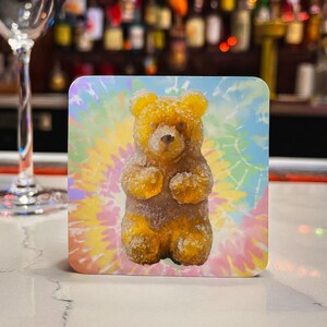 May include: A square drinks coaster with a gummy bear design. The gummy bear is golden with a sugared coating, set against a tie-dye background of blue, yellow, pink, and green. The coaster is on a white surface.