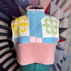 Pode incluir: Um colete acolchoado com gola alta e fecho de meio zíper. O colete apresenta um design de patchwork com padrões florais amarelos, verdes e azuis. A metade inferior é um material macio e rosa.