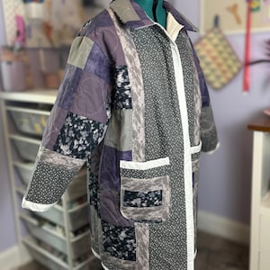 May include: A patchwork coat in shades of gray, purple, and black, displayed on a mannequin. The coat features a variety of fabric patterns, including polka dots and floral prints. It has a collar, pockets, and a white trim along the edges.