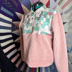 May include: A pink fleece pullover with a quilted patchwork design on the upper chest and collar. The patchwork features squares of various floral and patterned fabrics in shades of green, pink, and blue. The pullover has a half-zip closure.