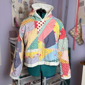 May include: A colorful patchwork hoodie with a white hood and cuffs. The jacket features a variety of patterned fabrics in shades of pink, yellow, blue, and green. Small pink pom-poms are sewn onto the seams. The jacket is displayed on a mannequin.