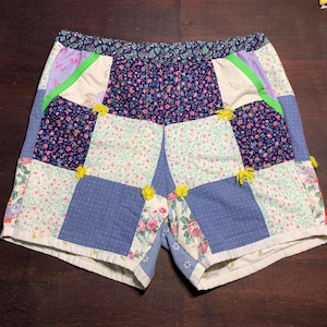Puede incluir: Pantalones cortos de patchwork hechos con diferentes telas florales en tonos azules, rosas, verdes y blancos. Los pantalones cortos tienen una cintura blanca y pompones amarillos en las costuras.