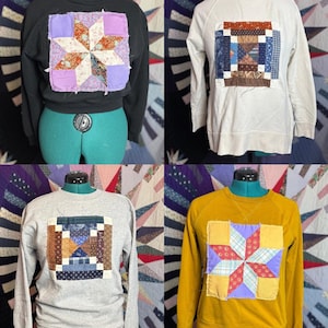 Puede incluir: Cuatro sudaderas en negro, blanco, gris y amarillo mostaza, cada una con un diseño de patchwork único. El patchwork incluye patrones de estrellas y geométricos en varios colores y telas. Las sudaderas se exhiben en maniquíes.