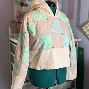 May include: A patchwork hoodie in pastel colors, including pink, green, and blue squares. The hoodie has a drawstring hood and is displayed on a mannequin. The design features small pink knots at the seams, adding a decorative touch to the quilted fabric.