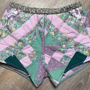 May include: Quilted shorts with a patchwork design. The shorts feature a floral pattern in shades of pink, green, and purple. The waistband is a dark green floral print. The shorts are made of a soft, quilted fabric.