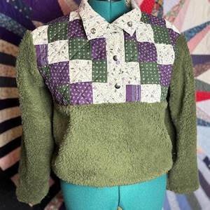 May include: Olive green fleece jacket with a quilted patchwork front featuring floral and geometric patterns in purple, green, and cream. The jacket has a collared neckline and button closure, offering a unique blend of textures and colors.