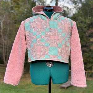 May include: Cropped hoodie with a patchwork design in pink and teal. The sleeves are a soft, fuzzy pink, and the hood is lined with a matching pink fabric. The garment is displayed on a green dress form.