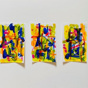 May include: Three abstract paintings on white paper. Each painting features a colourful, geometric design with bold strokes of blue, yellow, green, pink, and orange paint.