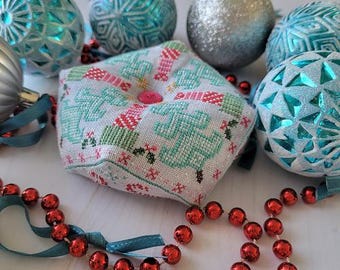 Op de afbeelding: Een witte kruissteek naaldhouder met een groen, roze en blauw kerstontwerp. De naaldhouder is omringd door blauwe en zilveren ornamenten.