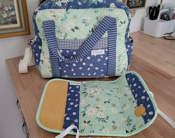 May include: A quilted fabric bag featuring a light mint green and dark blue floral pattern with white daisies. The bag includes dark blue gingham accents, two top handles, and a matching detachable folding mat with a solid mustard yellow panel.