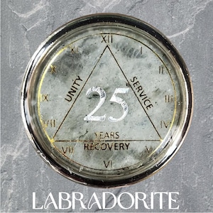 May include: A round medallion featuring a polished labradorite stone inlay with a silver-colored metal rim. The face displays a gold-printed triangle containing the words UNITY, SERVICE, 25 YEARS RECOVERY, and Roman numerals I through XII.