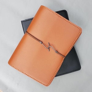 May include: Two leather-bound journals, one brown and one black, are stacked on top of each other. The brown journal has a black cord with a metal hook closure.