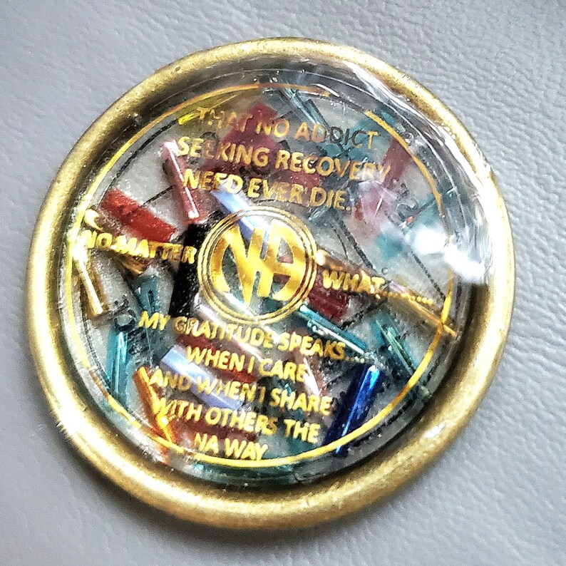 May include: A round, gold-colored medallion with a clear, domed center. Inside, colorful shards are visible. The text reads: "THAT NO ADDICT SEEKING RECOVERY NEED EVER DIE." The NA logo is in the center. Additional text includes "NO MATTER WHAT" and "MY GRATITUDE SPEAKS."
