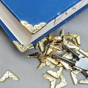 May include: A blue book with gold-colored corner protectors. The decorative metal corners are ornate and scattered around the book. A pair of pliers is also visible.