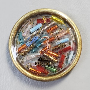 May include: A round, gold-toned pin with a clear resin center. Inside, colorful beads and text are visible, including the words "UNITY", "SERVICE", and "RECOVERY". The pin has a vintage aesthetic.