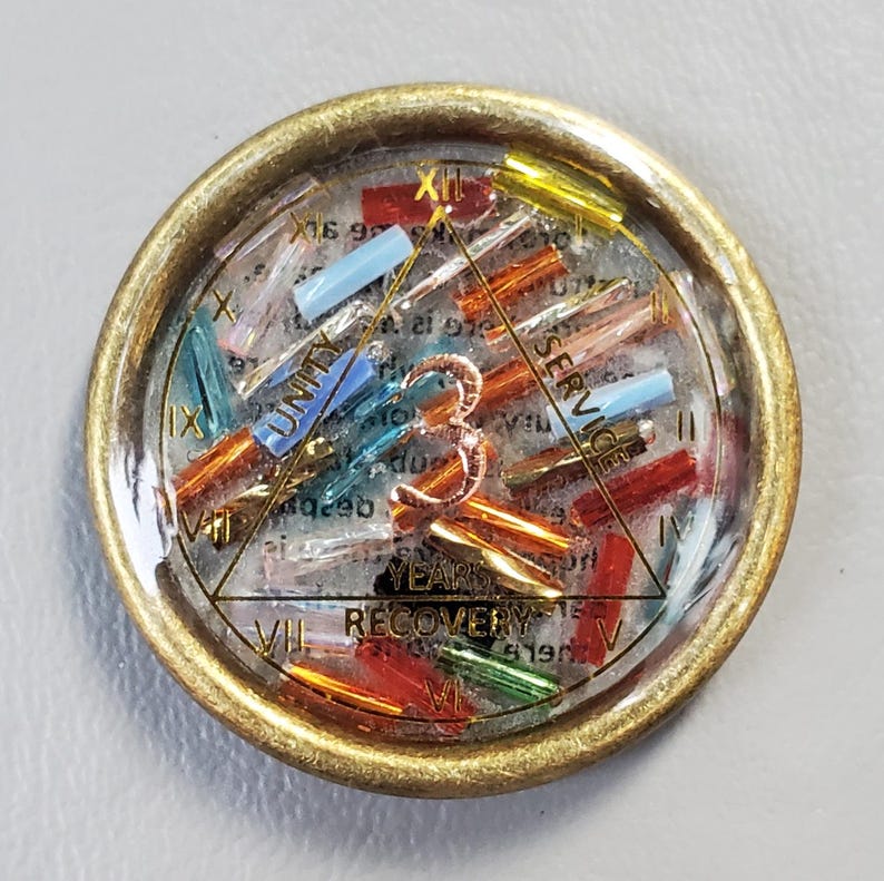 May include: A round, gold-toned pin with a clear resin center. Inside, colorful beads and text are visible, including the words "UNITY", "SERVICE", and "RECOVERY". The pin has a vintage aesthetic.