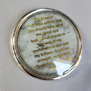 May include: A circular silver-toned token featuring a light gray, speckled stone-like center. Gold text is printed on the surface, reading: My Creator I am now willing that you should have all of me, good and bad. Which stands in the way of my usefulness to you and my fellows. Grant me strength, as I go out from here to do your bidding.
