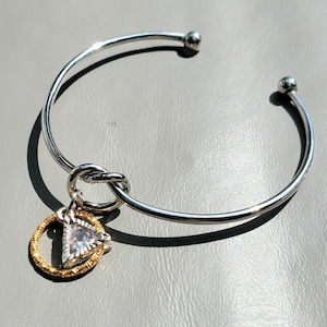 Puede incluir: Un brazalete de plata con un detalle de nudo y un dije dorado con una piedra triangular transparente.