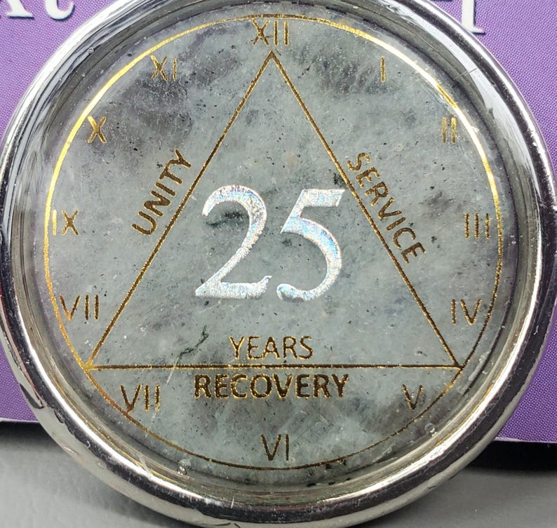 May include: A circular silver-toned medallion with a gray speckled stone center. It features a gold triangle with the words UNITY, SERVICE, and RECOVERY. The number 25 is centered in silver. Roman numerals I through XII are printed around the edge.