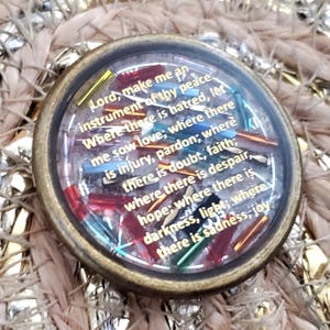 May include: A round, antique-style pendant with a clear glass center, featuring colorful accents and text. The text reads "Lord, make me an instrument of thy peace..." The pendant has a bronze-colored metal frame and is set against a woven background.