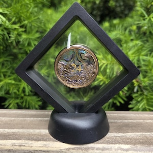 May include: A round, iridescent coin with the number 24 in gold, encased in a clear display case with a black frame. The display case is supported by a black base, set against a backdrop of green foliage.