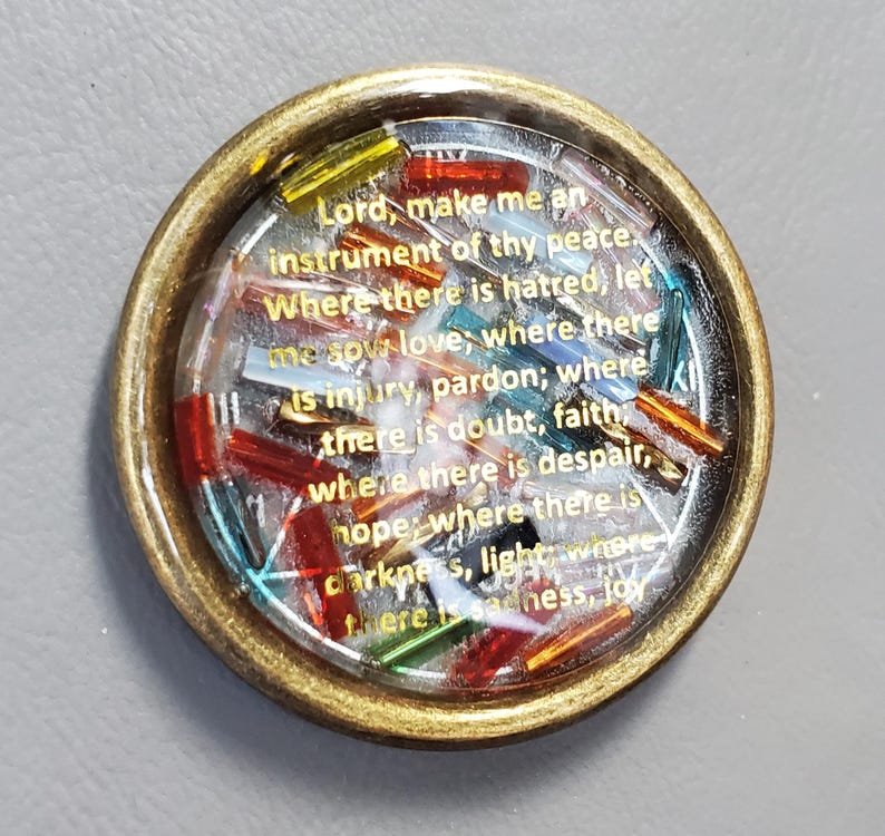 May include: A round, gold-toned pendant with a clear resin center. Inside, colorful glass beads and gold text read "Lord, make me an instrument of thy peace." The pendant has a vintage aesthetic.