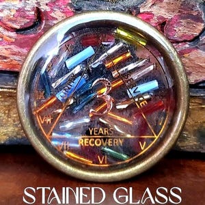May include: A round, stained glass piece with a bronze-colored metal frame. The glass features colorful, rectangular pieces and the words "UNITY" and "YEARS RECOVERY". Roman numerals mark the hours around the edge. The text "STAINED GLASS" is at the bottom.