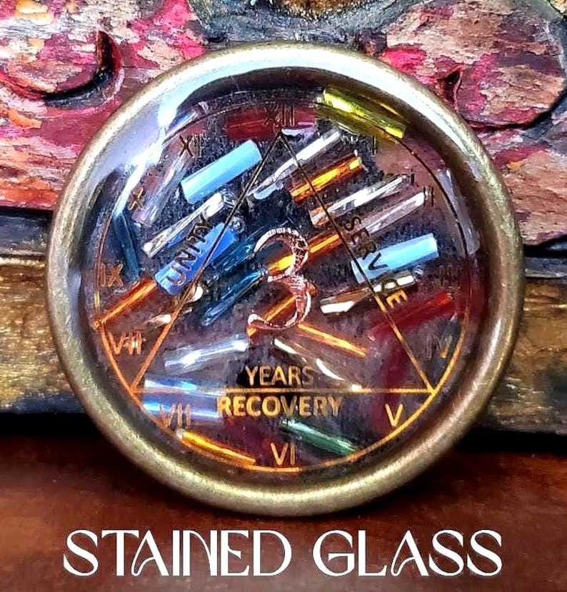 May include: A round, stained glass piece with a bronze-colored metal frame. The glass features colorful, rectangular pieces and the words "UNITY" and "YEARS RECOVERY". Roman numerals mark the hours around the edge. The text "STAINED GLASS" is at the bottom.