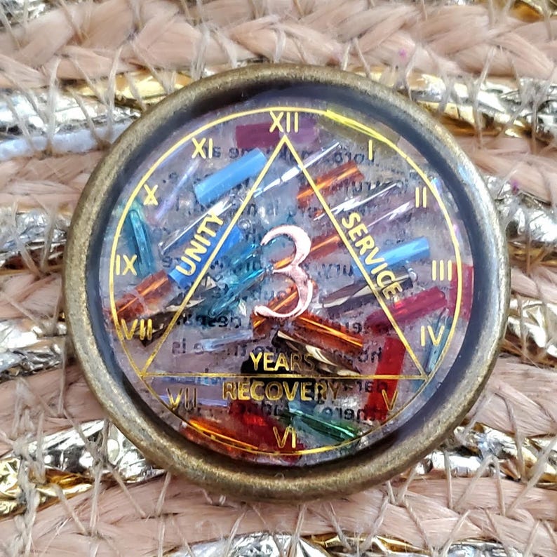May include: A round, bronze-colored medallion with a clear center filled with colorful, small glass pieces. The number "3" is prominent, with the words "UNITY" and "SERVICE" on a triangle. The words "YEARS" and "RECOVERY" are also visible.