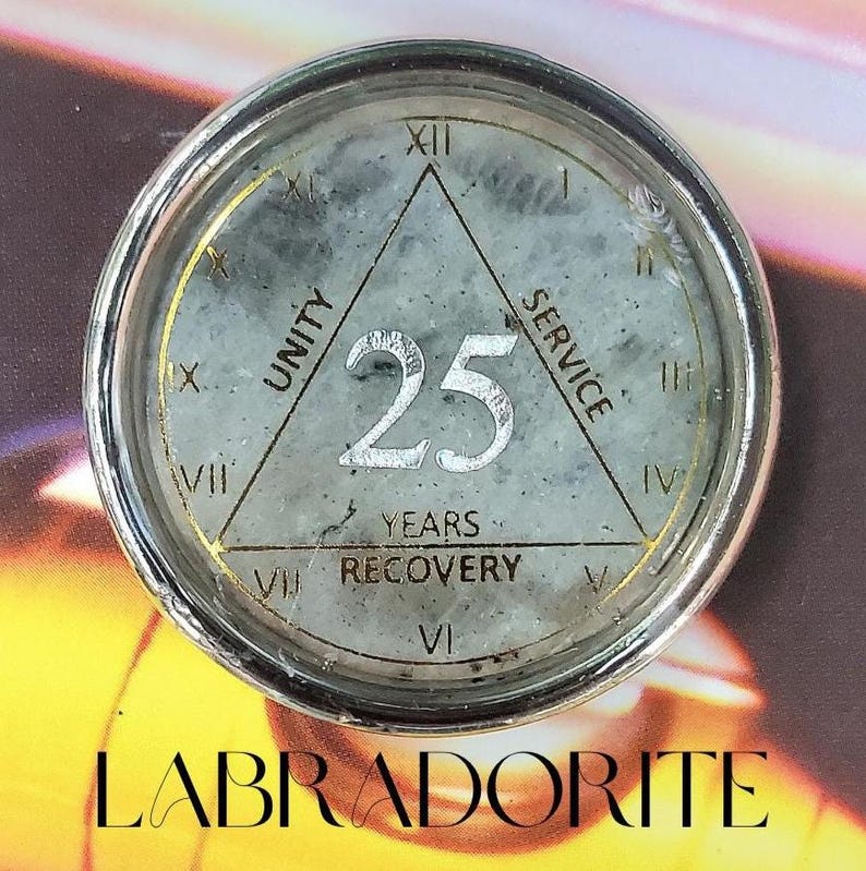 May include: A round, silver-toned medallion featuring a grey labradorite stone inlay. The center displays the number 25, surrounded by a triangle with the words UNITY, SERVICE, and RECOVERY. Roman numerals I through XII encircle the perimeter.