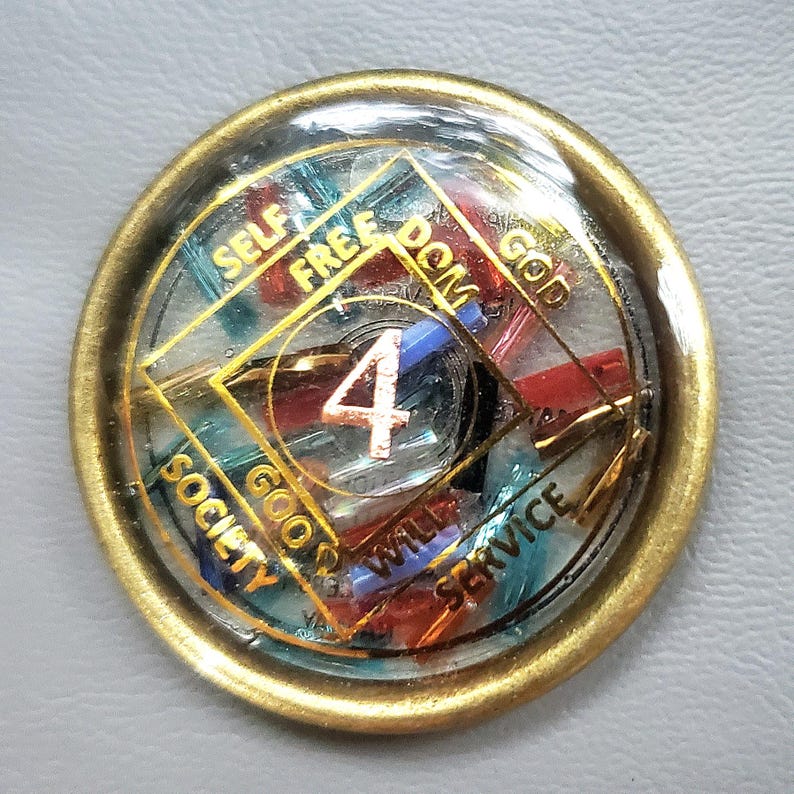 May include: A round, gold-colored medallion with a clear, resin-like center. The center features a gold-outlined diamond shape with the number "4" in the center. The words "SELF", "FREEDOM", "GOD", "SERVICE", "WILL", "GOOD", and "SOCIETY" are visible.