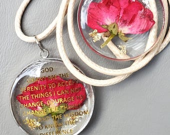 May include: A silver necklace with a round pendant featuring a pressed red flower and the text "GOD GRANT ME THE SERENITY TO ACCEPT THE THINGS I CAN NOT CHANGE, COURAGE TO CHANGE THE THINGS I CAN AND THE WISDOM TO KNOW THE DIFFERENCE".