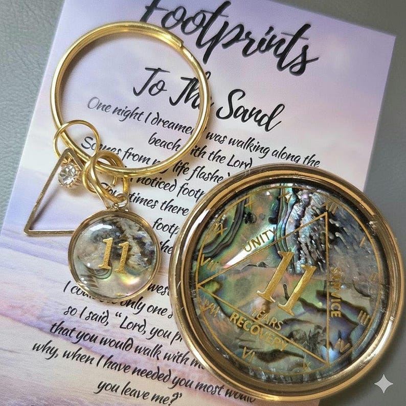 Peut inclure: Un porte-cl&eacute;s dor&eacute; avec un charm en forme de triangle et un charm rond avec le chiffre 11. Un m&eacute;daillon rond avec le chiffre 11 et les mots "Unity" et "Recovery" est &eacute;galement visible. L'arri&egrave;re-plan comprend du texte d'un po&egrave;me.
