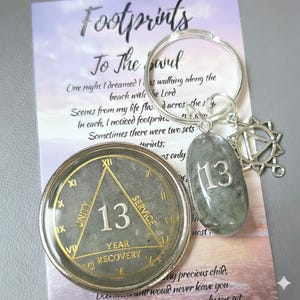 May include: A 13-year recovery medallion featuring a gold triangle with the words Unity, Service, and Recovery, alongside a silver keychain with a grey stone charm engraved with the number 13 and a star-shaped metal charm on a printed card.