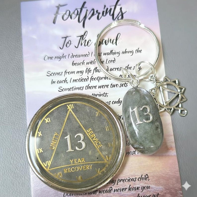 May include: A 13-year recovery medallion featuring a gold triangle with the words Unity, Service, and Recovery, alongside a silver keychain with a grey stone charm engraved with the number 13 and a star-shaped metal charm on a printed card.