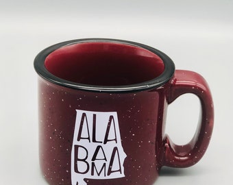 Puede incluir: Una taza de cerámica granate con borde y asa negros. La taza presenta un gráfico blanco del estado de Alabama con la palabra "ALABAMA" en negrita, sin serifa. La taza tiene un acabado moteado.