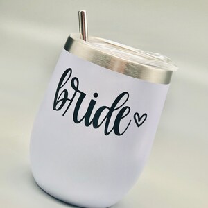 K&ouml;nnte beinhalten: Ein wei&szlig;er Edelstahlbecher mit einem schwarzen Vinyl-Aufkleber, auf dem "bride" mit einem kleinen Herzen steht. Der Becher hat einen Deckel und einen Strohhalm.