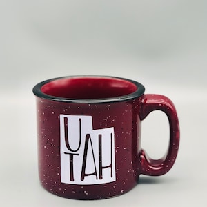 May include: A maroon ceramic mug with a speckled finish. The mug has a white outline of the state of Utah with the word "Utah" written inside the outline.