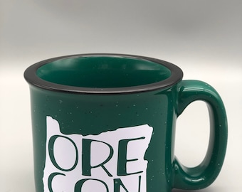Puede incluir: Taza de cerámica verde con borde negro y un contorno blanco del estado de Oregón con la palabra "OREGON" escrita en su interior.